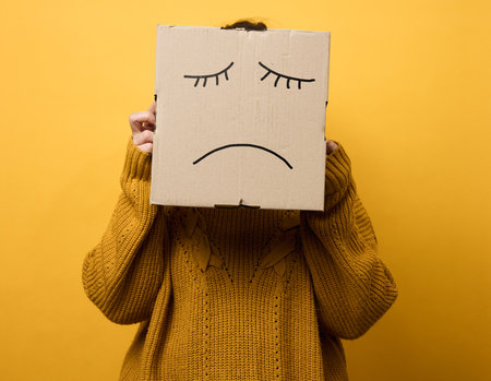 a woman in a brown sweater with a box on her head stands on a yellow background. Sad grimace, stress and depression concept. Mental healthの写真素材