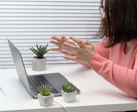 woman in a pink sweater sits at a table and looks at a laptop, makes a video call, gesturing with...