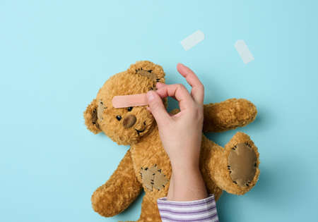 female hand holds a brown teddy bear and glues a medical adhesive plaster on a blue background, tram treatmentの写真素材