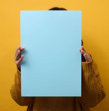 woman in an orange sweater holds a blank sheet of paper on a yellow background. Place for an inscription, advertisement, informationの写真素材