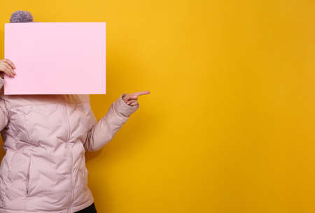 woman in pink winter jacket and hat holds blank pink sheet of paper on yellow background. Seasonal sale. Place for inscriptionの写真素材