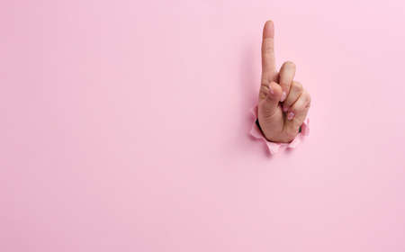 female hand sticking out of a torn hole in a pink paper background, attention gesture. Place for an inscriptionの写真素材
