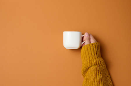 white ceramic cup in a female hand on orange background. drink and hand are raised up, coffee break.の写真素材