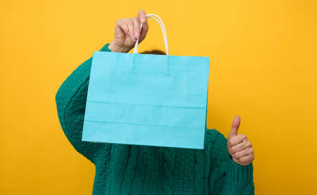 a woman in a sweater holds a paper bag on a yellow background and shows a like gesture with her...