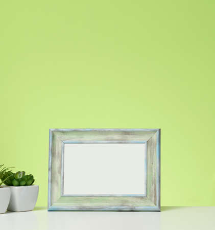 empty white wooden photo frame and flowerpots with plants on white table, green backgroundの写真素材