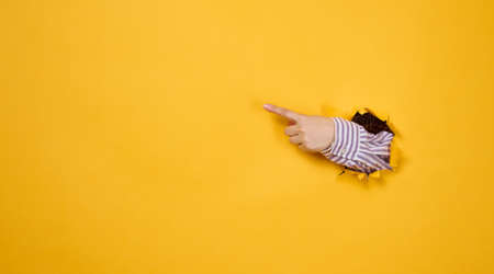female hand sticks out of a torn hole in a yellow paper background, part of the body points with the index finger to the side. Copy spaceの写真素材