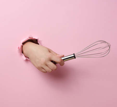 female hand holds a metal whisk for beating products on a pink background. A part of the body sticks out of a torn holeの写真素材