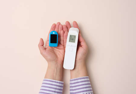 female hand holds plastic non-contact thermometer and oximeter on beige background