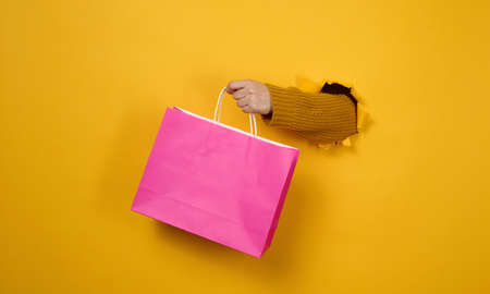 female hand with a pink paper shopping bag sticking out of a torn hole in a yellow paper background. Refusal from plastic, start of salesの写真素材
