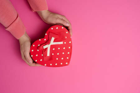 two female hands hold a red heart-shaped box on a pink background. Festive backdrop
