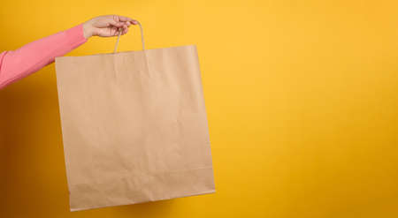 woman holding in hand brown blank craft paper bag for takeaway on yellow background. Packaging template mock up. Delivery service concept, bannerの写真素材