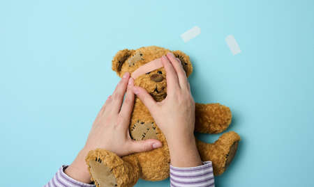female hand holds a brown teddy bear and glues a medical adhesive plaster on a blue background, tram treatmentの写真素材
