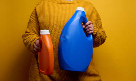 woman in a knitted sweater holds an orange and blue plastic bottles with liquid washing gel. Laundry and chores, yellow backgroundの写真素材