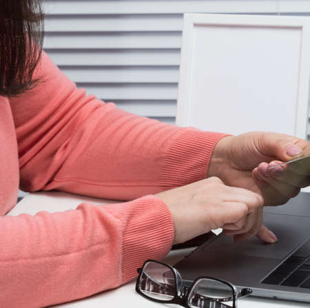 woman in a pink sweater makes purchases online with a laptop, a credit card in hand. A woman sits on...