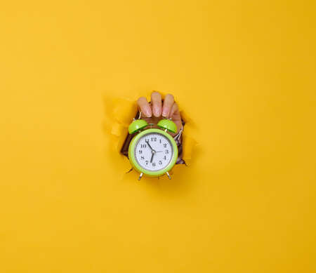 a woman's hand holds a round green alarm clock, the time is five minutes to seven in the morning. A part of the body is sticking out of a torn hole in a yellow paper background.の写真素材