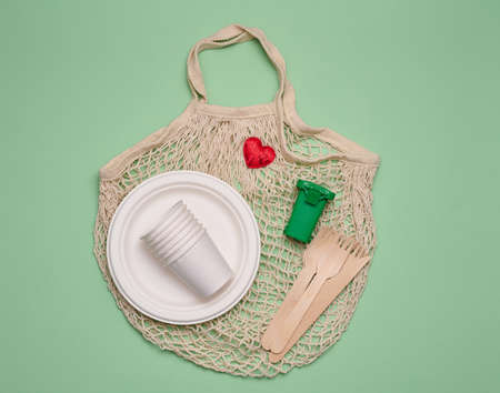 white cotton bag, paper cups and wooden forks and spoons on a green background. Recyclable waste, top viewの写真素材