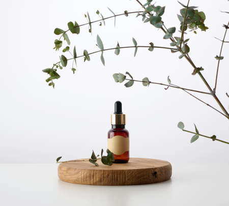 a glass brown bottle with a pipette on a wooden podium and a branch of eucalyptus, white background. Advertising and promotion of cosmetic productsの写真素材