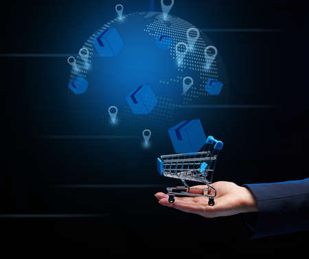 a hand holds a miniature shopping cart against the background of a holographic planet earth with parcel delivery points. international tradeの写真素材