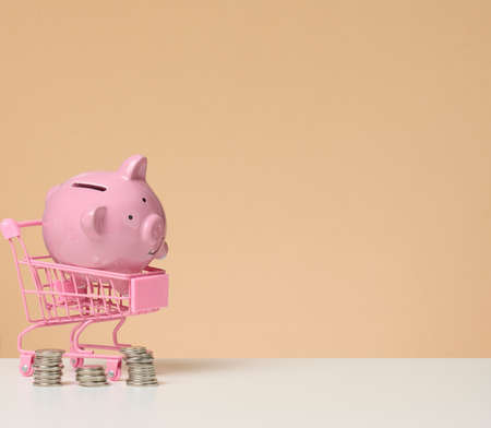 metal miniature shopping cart and pink piggy bank on a white table, brown background. Concept of saving budget, discounts, copy spaceの写真素材