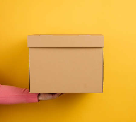 female hand holding closed brown cardboard box on yellow background, deliveryの写真素材