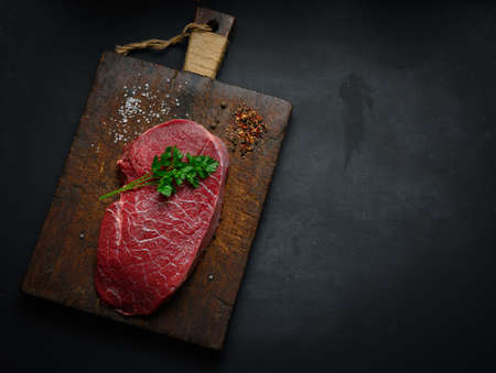Raw beef tenderloin lies on a cutting board and spices for cooking on a black table, top view