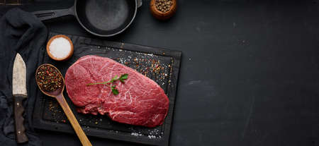 Raw beef tenderloin lies on a cutting board and spices for cooking on a black table, top view