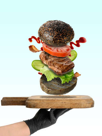 A hand holds a wooden cutting board and flying hamburger ingredients. Black rye bun with sesame, slice of tomato and cucumber, fried piece of meatの写真素材