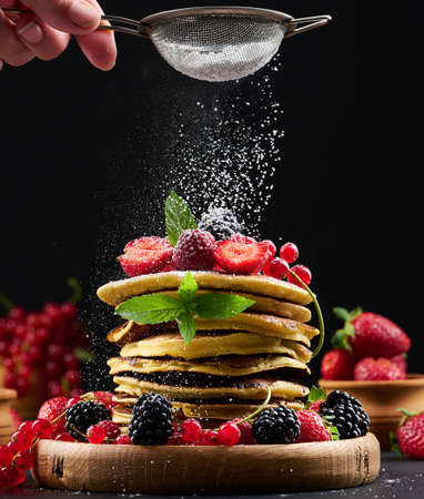 A stack of pancakes with fresh fruit sprinkled with powdered sugar on a black backgroundの写真素材