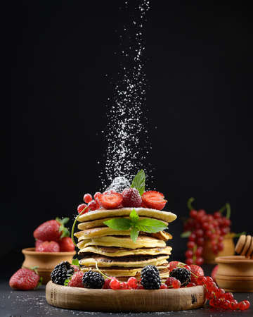 A stack of pancakes with fresh fruit sprinkled with powdered sugar on a black backgroundの写真素材