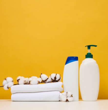 White plastic bottles for gel and shampoo and rolled white terry towels on a white table, yellow...