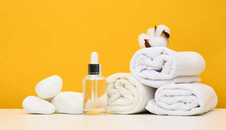 Glass bottle with a pipette and twisted white terry towels on the table, yellow backgroundの写真素材