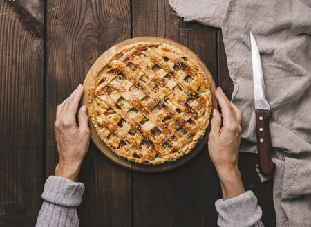Two female hands hold a round baked pie with apple filling on a wooden board, brown table. View from aboveの写真素材