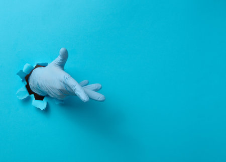 Hand in a blue medical glove is sticking out of a torn hole in a blue paper background. Part of the body indicates the direction, copy spaceの写真素材