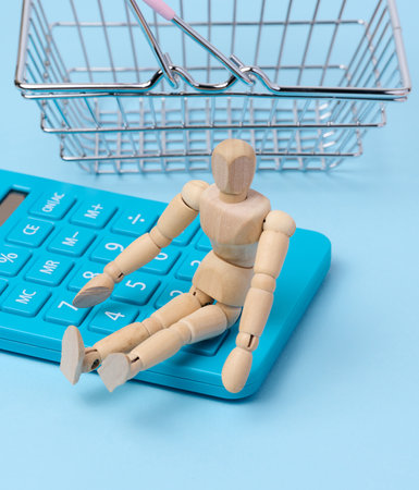 A wooden puppet and a blue calculator, behind a miniature shopping basketの写真素材