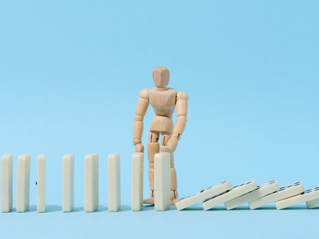 A wooden puppet toy holds back a falling domino on a blue background, representing the concept of a strong personalityの写真素材