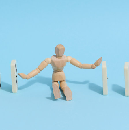 A wooden puppet toy holds back a falling domino on a blue background, representing the concept of a strong personalityの写真素材