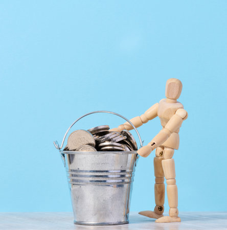 A wooden mannequin and coins in a miniature bucket on a blue background, a concept of high income, subsidyの写真素材
