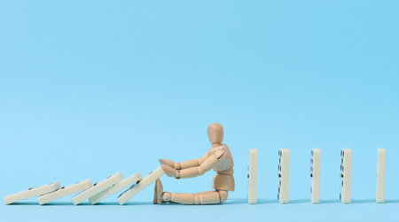 A wooden puppet toy holds back a falling domino on a blue background, representing the concept of a strong personalityの写真素材