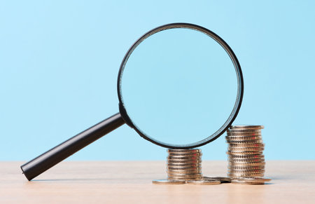 A stack of metal coins and a plastic magnifier on a blue background. The concept of increasing taxes, subsidiesの写真素材