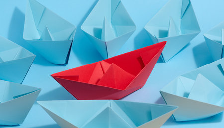 A red paper boat stands in front of a group of blue paper boats, a confrontationの写真素材
