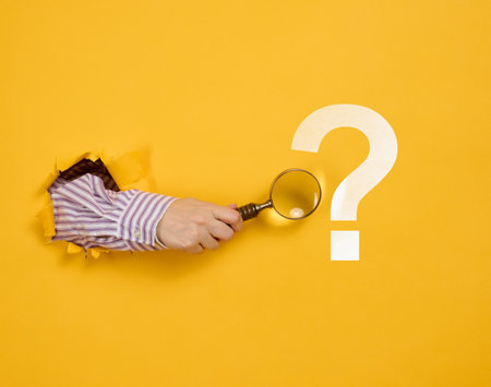 Female hand holds a magnifying glass on a yellow background, part of the body sticks out of a torn hole in a paper background. Business concept, finding information, answering questionsの写真素材