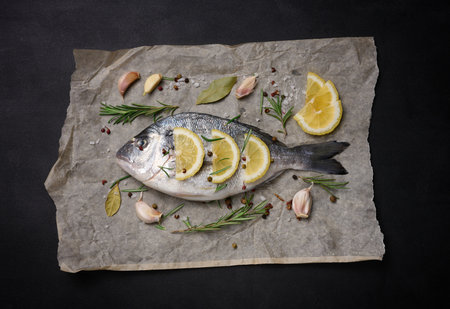 Raw whole dorado fish on brown parchment paper and spices for cooking, top view on black tableの写真素材