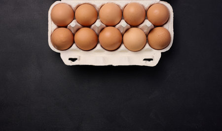 Raw brown chicken eggs in a paper cardboard container on a black background, top view