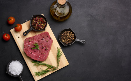 Raw piece of beef with spices pepper, rosemary sprig, salt and olive oil on a wooden board, black...