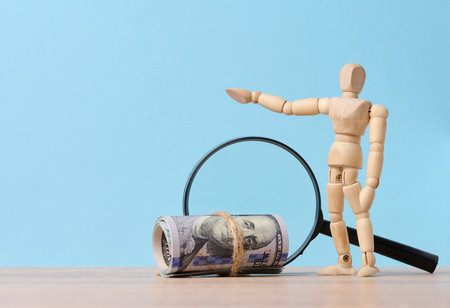 Wooden mannequin, magnifying glass and tied paper american dollars on a blue background, the concept of income growth, searching for a profitable offerの写真素材