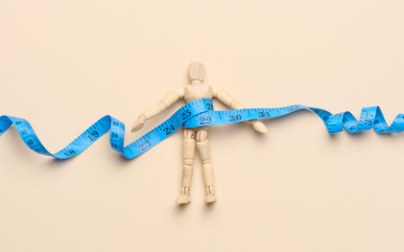 Wooden mannequin wrapped with blue measuring tape on a beige background, weight loss conceptの写真素材