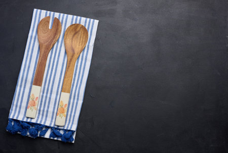 Two wooden spoons on a striped white textile kitchen towel on a black background, top view