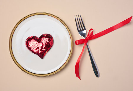 Red heart on a round white plate and fork on a beige background, top view
