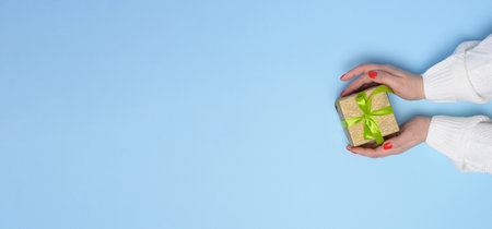 Female hands holding a gift box with a green ribbon on a blue background. View from above, copy...