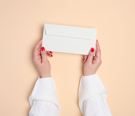Two female hands holding a white rectangular envelope on a beige background, top viewの写真素材